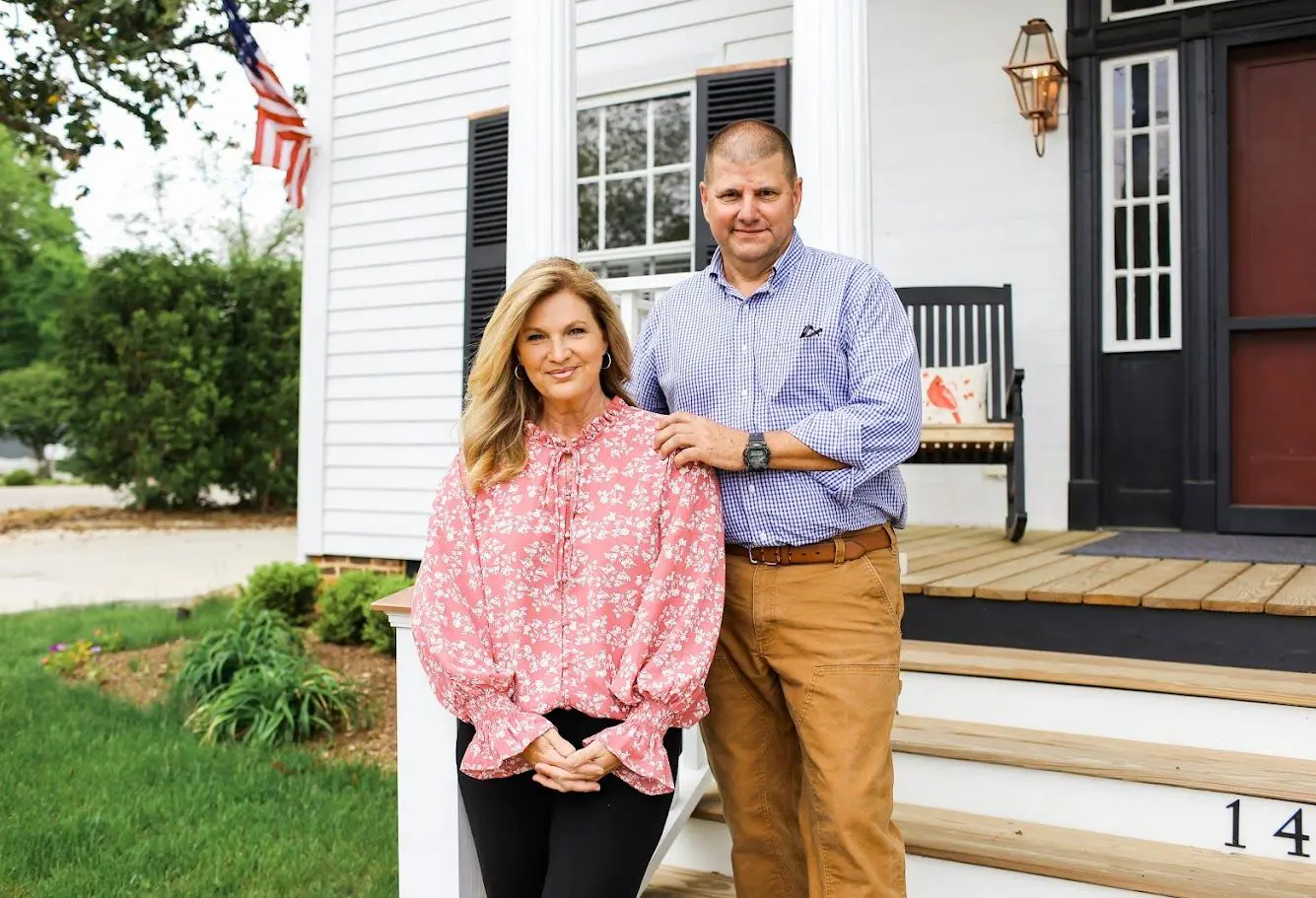 A smiling couple standing outside a white house with a porch.
