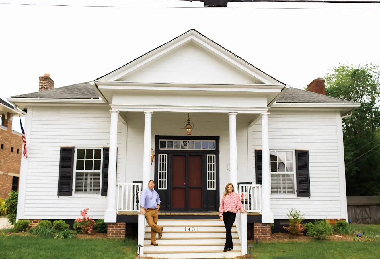 Two people stand on the steps of a white house with columns.