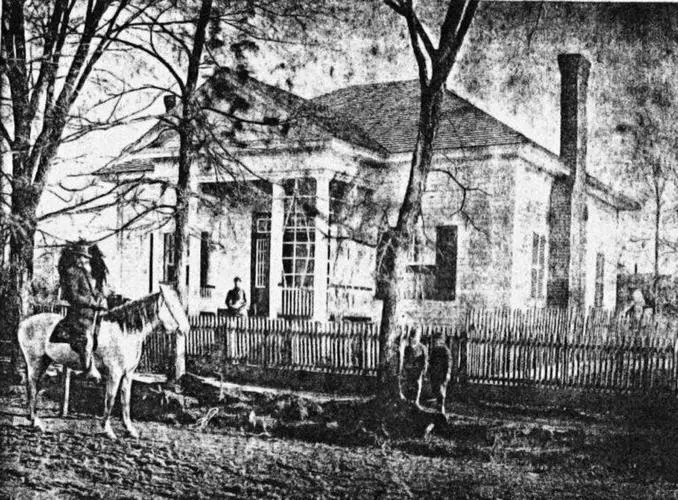 Black and white photo of a horse and rider outside a historic farmhouse.