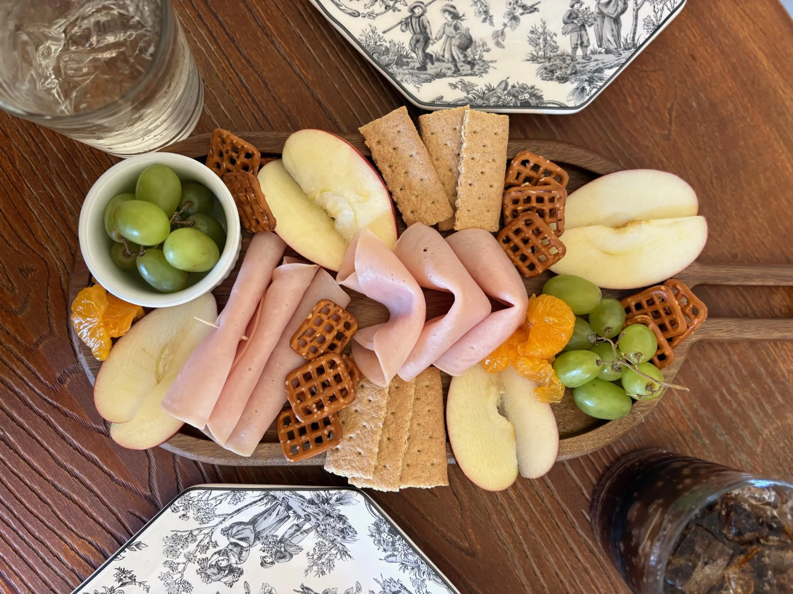 A wooden board with sliced turkey, apples, crackers, and grapes.